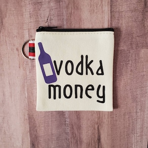 May include: A small, white canvas coin purse with a black zipper and a black and red zipper pull. The purse has a purple silhouette of a liquor bottle and the text "vodka money" printed on it.