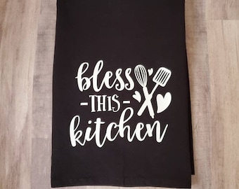 Puede incluir: Toalla de cocina negra con texto blanco que dice "Bless this kitchen" con un batidor, una espátula y un gráfico en forma de corazón.