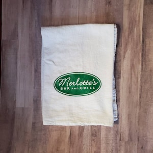 May include: White kitchen towel with a green oval logo that says "Merlotte's Bar and Grill".