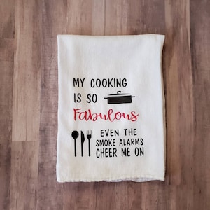 May include: White kitchen towel with black text that reads "My cooking is so fabulous even the smoke alarms cheer me on". The text is in a black font with a red outline. There is a black silhouette of a pot and a spoon, fork, and knife.