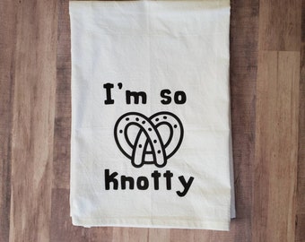 May include: White kitchen towel with black text that reads "I'm so knotty" and a black pretzel graphic.