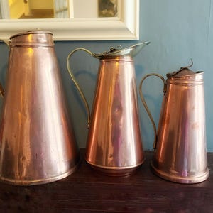 May include: Three copper jugs of different sizes, each with a brass handle and lid. They have a conical shape and are displayed on a dark wooden surface. The background features a light blue wall and a white-framed mirror.