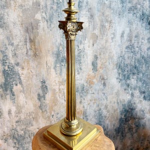 May include: A tall, gold-coloured decorative pillar with a square base and ornate capital. The pillar is made of metal and has a fluted shaft. The capital features detailed carvings. The pillar is displayed on a wooden surface against a textured wall.