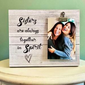 May include: A wooden photo frame with the words "Sisters are always together in Spirit" printed on it. The frame holds a photo of two smiling women. A metal clip holds the photo in place. The frame is sitting on a white nightstand.