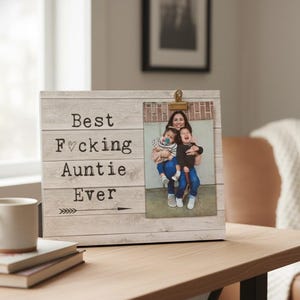 May include: A rectangular wooden photo frame with a light gray wood grain finish. The left side features the black text "Best F*cking Auntie Ever" with a heart symbol and an arrow. A gold metal clip holds a vertical photograph of three people.