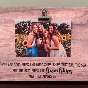May include: A wooden photo frame with a pinkish-brown finish, holding a photograph of four smiling women in front of sunflowers. The frame has a metal clip and the text: "THERE ARE GOOD SHIPS AND WOOD SHIPS, SHIPS THAT SAIL THE SEA BUT THE BEST SHIPS ARE friendships MAY THEY ALWAYS BE."