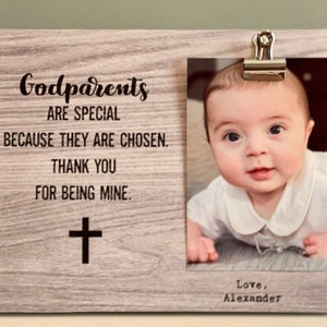 May include: A wooden photo frame with a baby's picture clipped to the right side. The frame has text that reads "Godparents are special because they are chosen. Thank you for being mine." A cross is below the text.
