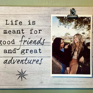 May include: A wooden photo frame with a printed message that reads "Life is meant for good friends and great adventures." The frame holds a photograph of two smiling people. A metal clip secures the photo.