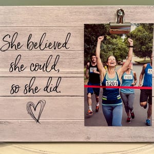 May include: Wooden picture frame with a photo of a marathon runner crossing the finish line. The frame has the quote "She believed she could, so she did" and a heart drawing. The frame is light gray.