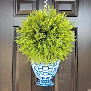 May include: A decorative door hanging featuring a blue and white vase design filled with bright green faux foliage. The vase has a classic pattern and is suspended from a black door.