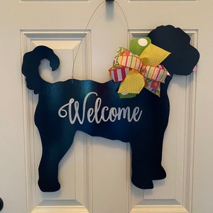 May include: Black silhouette of a dog with a yellow, pink, and green bow, hanging on a white door with the word "Welcome" written in white.