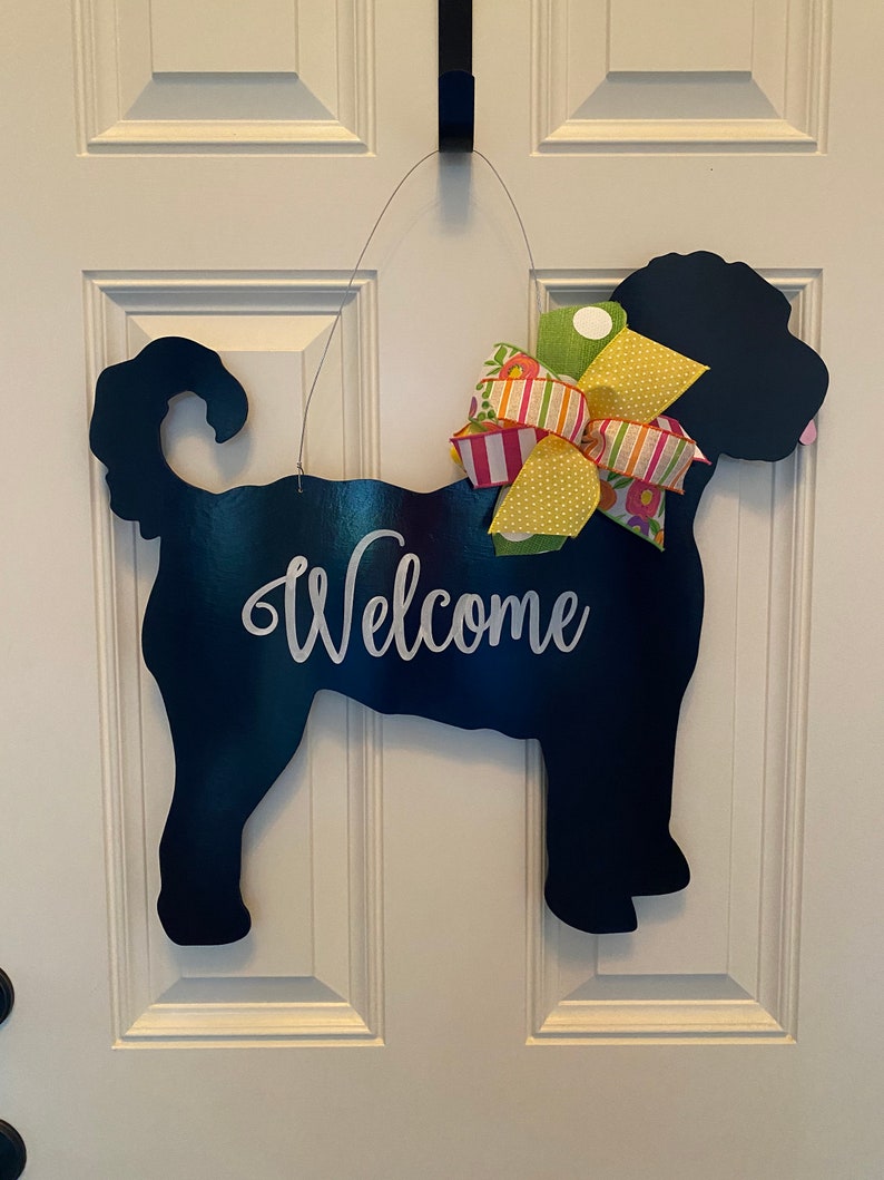May include: Black silhouette of a dog with a yellow, pink, and green bow, hanging on a white door with the word "Welcome" written in white.