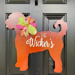 May include: A wooden dog silhouette door hanger painted in shades of orange and red. The dog is wearing a pink, green, and white bow. The text "THE Wicker's" is written on the dog's body.