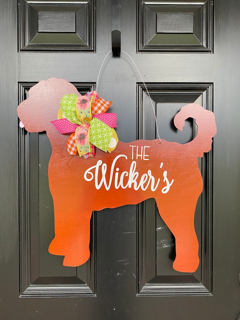 May include: A wooden dog silhouette door hanger painted in shades of orange and red. The dog is wearing a pink, green, and white bow. The text "THE Wicker's" is written on the dog's body.