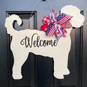 May include: A white wooden dog silhouette door hanger with the word "Welcome" written on it. The dog is decorated with a red, white, and blue bow.