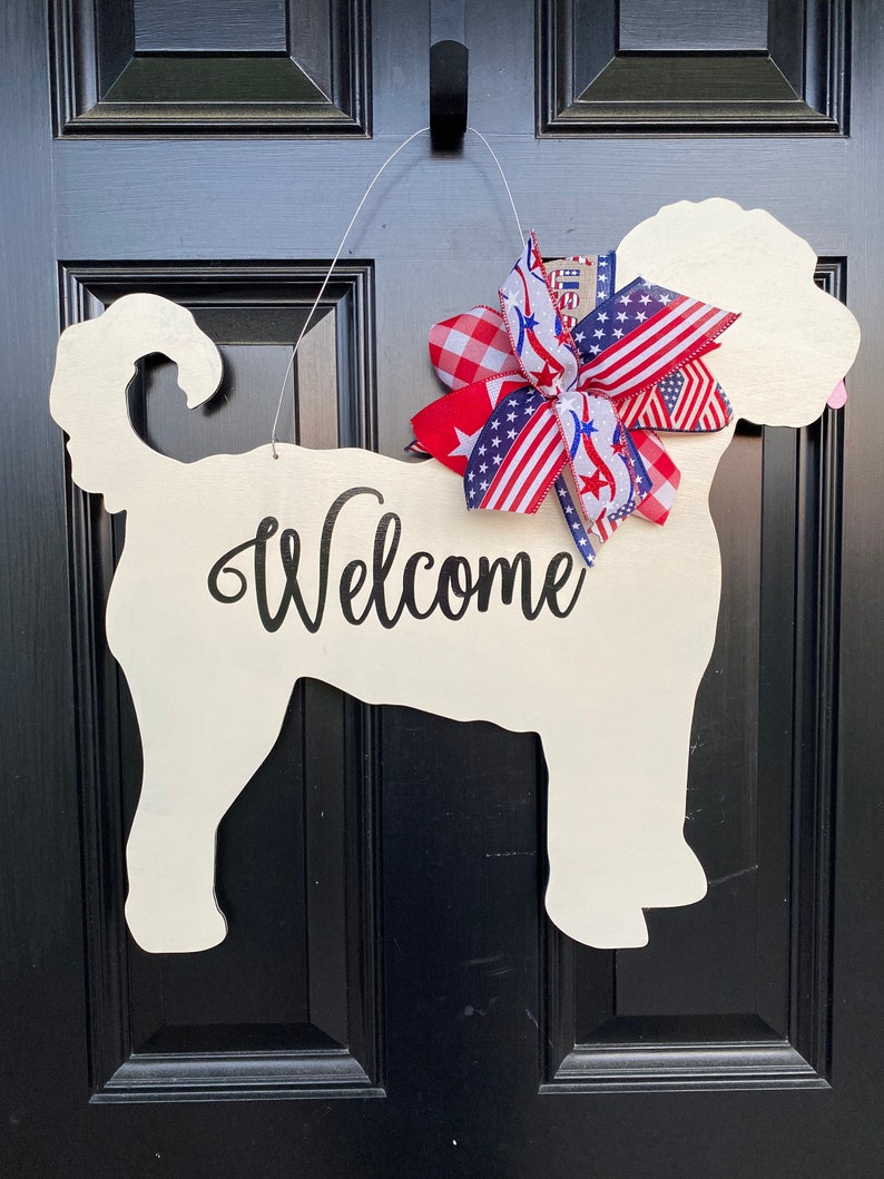 May include: A white wooden dog silhouette door hanger with the word "Welcome" written on it. The dog is decorated with a red, white, and blue bow.