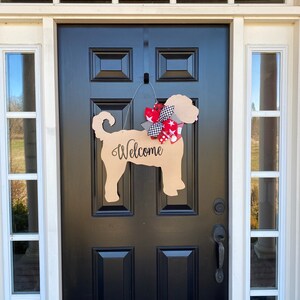 May include: A wooden dog-shaped welcome sign with a red, white, and black bow hanging on a black front door. The sign says "Welcome" in black lettering.