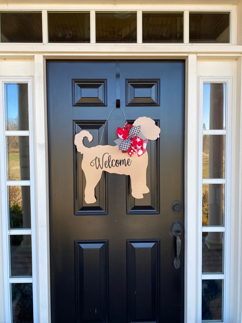 May include: A wooden dog-shaped welcome sign with a red, white, and black bow hanging on a black front door. The sign says "Welcome" in black lettering.