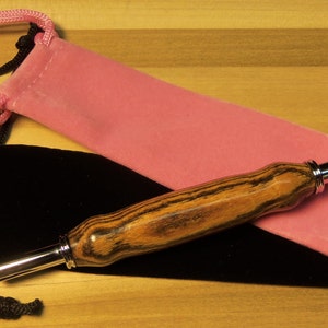 Pode incluir: Uma caneta de madeira com ponta e detalhes prateados repousa sobre uma bolsa de veludo preta e rosa. A caneta tem um padr&atilde;o listrado em tons de marrom e preto. A bolsa rosa est&aacute; aberta e a bolsa preta est&aacute; fechada.