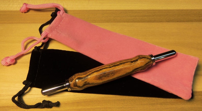 Pode incluir: Uma caneta de madeira com ponta e detalhes prateados repousa sobre uma bolsa de veludo preta e rosa. A caneta tem um padr&atilde;o listrado em tons de marrom e preto. A bolsa rosa est&aacute; aberta e a bolsa preta est&aacute; fechada.