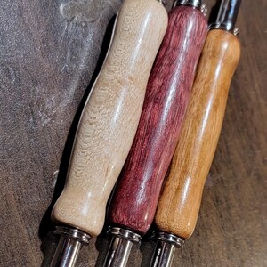 Pode incluir: Quatro rasgadores de costura com cabos de madeira em v&aacute;rias cores: bege claro, vermelho escuro, castanho m&eacute;dio e castanho claro. Cada um tem uma l&acirc;mina de metal prateado com uma ponta vermelha. As ferramentas est&atilde;o dispostas diagonalmente sobre uma superf&iacute;cie castanha escura.