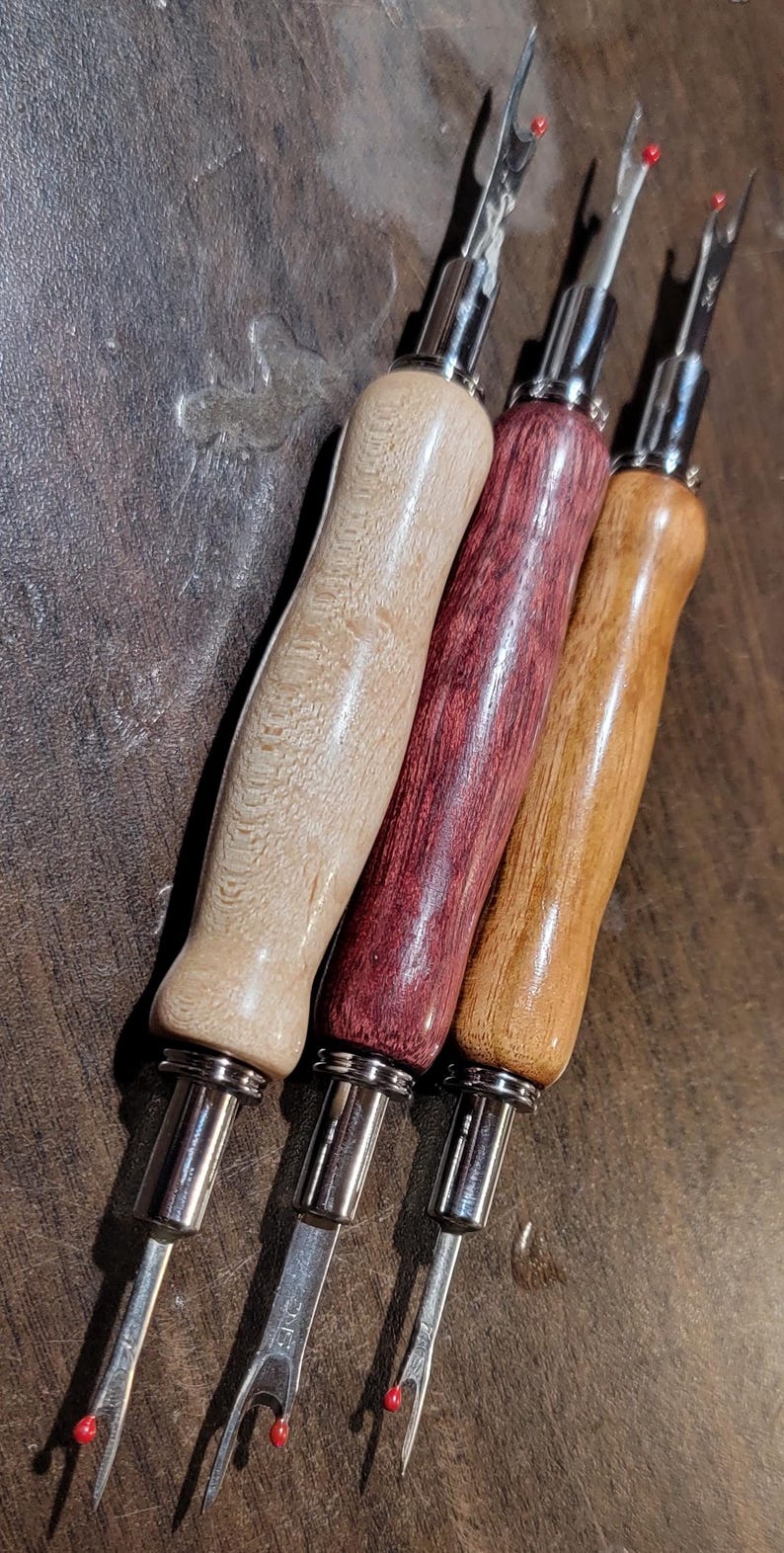 Pode incluir: Quatro rasgadores de costura com cabos de madeira em v&aacute;rias cores: bege claro, vermelho escuro, castanho m&eacute;dio e castanho claro. Cada um tem uma l&acirc;mina de metal prateado com uma ponta vermelha. As ferramentas est&atilde;o dispostas diagonalmente sobre uma superf&iacute;cie castanha escura.