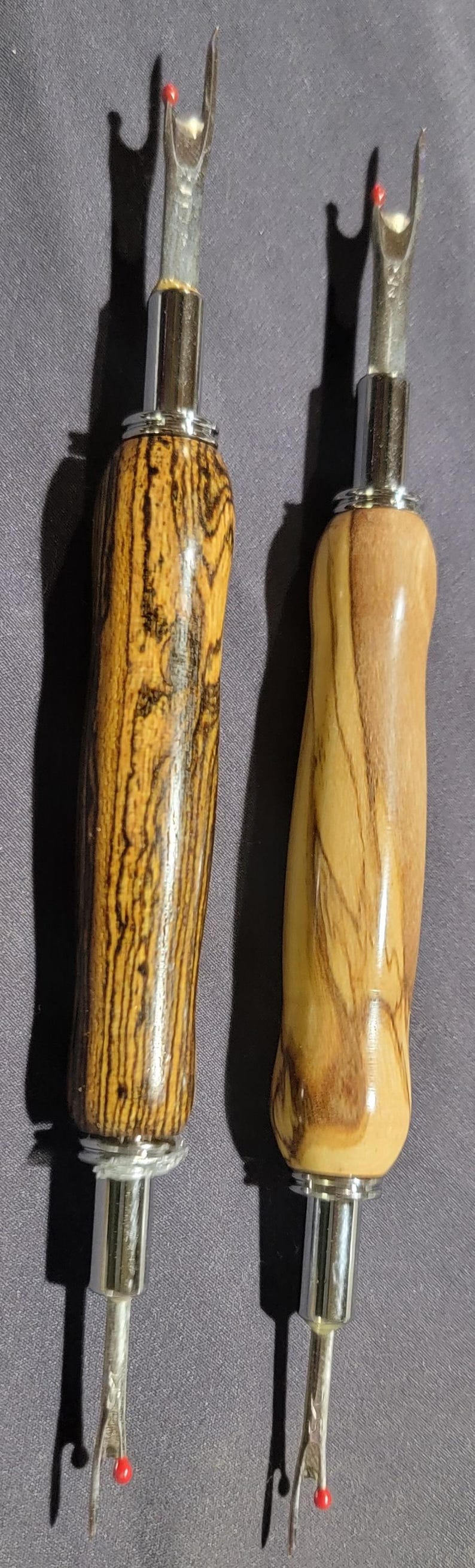 Pode incluir: Dois abre-costuras com cabos de madeira e l&acirc;minas de metal prateado. Uma pega &eacute; castanha escura com riscas pretas, a outra &eacute; castanha clara com riscas castanhas mais escuras. Cada um tem uma bola vermelha na ponta.