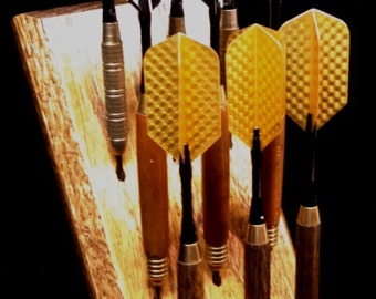 May include: A wooden dartboard holder with six darts. The darts have green, black and white, and gold fletching.