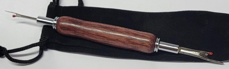Pode incluir: Um rasgador de costura com cabo de madeira e detalhes em metal prateado, em um saco preto. O cabo &eacute; de cor marrom escuro, e o rasgador tem uma ponta com uma pequena conta vermelha.