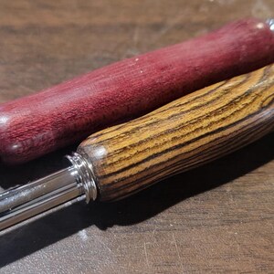Pode incluir: Duas ferramentas de madeira com pontas de metal. Uma &eacute; de cor vermelho escuro e a outra castanho claro com gr&atilde;o de madeira. As ferramentas s&atilde;o provavelmente para um artesanato ou passatempo, como tric&ocirc; ou croch&ecirc;.