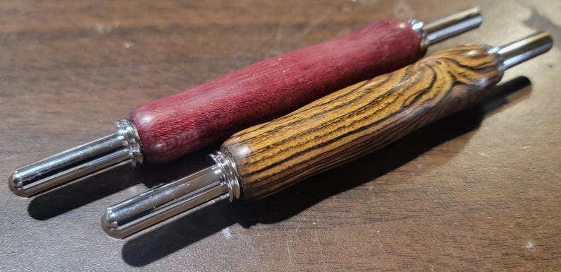 Pode incluir: Duas ferramentas de madeira com pontas de metal. Uma &eacute; de cor vermelho escuro e a outra castanho claro com gr&atilde;o de madeira. As ferramentas s&atilde;o provavelmente para um artesanato ou passatempo, como tric&ocirc; ou croch&ecirc;.