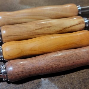 Pode incluir: Quatro agulhas de croch&ecirc; de madeira com extremidades de metal polido. Os cabos s&atilde;o feitos de diferentes tipos de madeira, variando de castanho claro a castanho escuro. As agulhas est&atilde;o dispostas em paralelo.