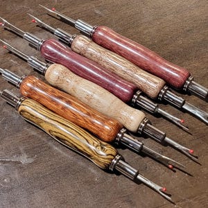 May include: A collection of seam rippers with wooden handles in various colors, including dark red, brown, and tan. Each tool has a silver metal shaft and a sharp blade with a red safety ball. The tools are arranged diagonally on a brown surface.