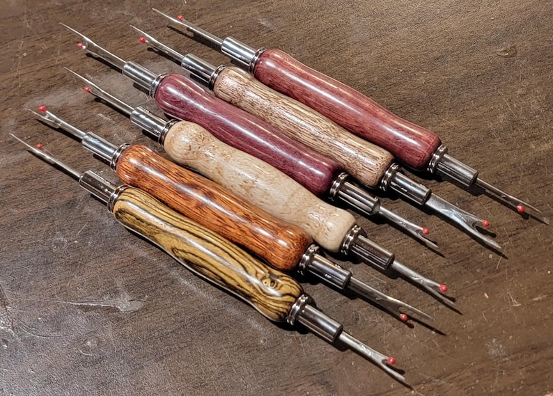 Pode incluir: Uma cole&ccedil;&atilde;o de desfibradores de costura com cabos de madeira em v&aacute;rias cores, incluindo vermelho escuro, castanho e bege. Cada ferramenta tem um eixo de metal prateado e uma l&acirc;mina afiada com uma bola de seguran&ccedil;a vermelha. As ferramentas est&atilde;o dispostas diagonalmente sobre uma superf&iacute;cie castanha.