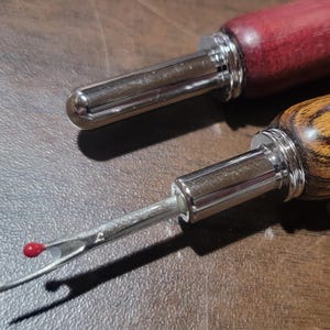 Pode incluir: Duas ferramentas de artesanato em couro com cabos de madeira e pontas de metal. Uma pega &eacute; de cor vermelho escuro, e a outra &eacute; castanho claro com um padr&atilde;o de gr&atilde;o de madeira. As pontas de metal t&ecirc;m um acabamento prateado, e uma tem uma pequena conta vermelha.