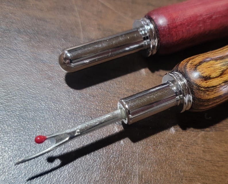 Pode incluir: Duas ferramentas de artesanato em couro com cabos de madeira e pontas de metal. Uma pega &eacute; de cor vermelho escuro, e a outra &eacute; castanho claro com um padr&atilde;o de gr&atilde;o de madeira. As pontas de metal t&ecirc;m um acabamento prateado, e uma tem uma pequena conta vermelha.