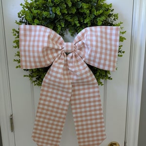 May include: A decorative wreath with a lush green base and a large, peach and white gingham bow. The bow is centered on the wreath, creating a charming and inviting aesthetic. Perfect for seasonal home decor.