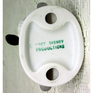 Puede incluir: Base de figura de cer&aacute;mica blanca con dos agujeros y soportes grises. El texto "WALT DISNEY PRODUCTIONS" est&aacute; impreso en verde. La figura tiene una forma redondeada con un asa curva.