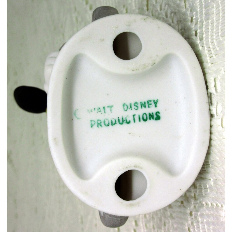 Puede incluir: Base de figura de cer&aacute;mica blanca con dos agujeros y soportes grises. El texto "WALT DISNEY PRODUCTIONS" est&aacute; impreso en verde. La figura tiene una forma redondeada con un asa curva.