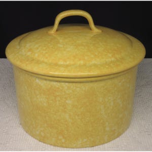 May include: A yellow ceramic casserole dish with a lid and handle. The dish has a speckled pattern and a rounded shape. The lid has a curved handle for easy lifting. The dish is designed for cooking and serving.