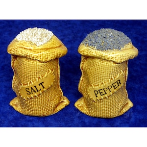 May include: A pair of ceramic salt and pepper shakers shaped like burlap sacks. The salt shaker is filled with white granules and labeled "SALT." The pepper shaker is filled with dark granules and labeled "PEPPER."