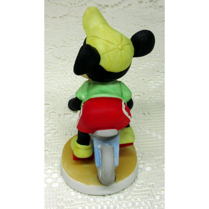 Puede incluir: Una figura de cer&aacute;mica de un rat&oacute;n de dibujos animados con una camisa verde, pantalones cortos rojos y una gorra amarilla, montando una motocicleta azul claro. La figura est&aacute; sobre una base redonda con una combinaci&oacute;n de colores beige y blanco.