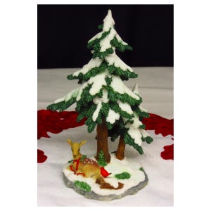 May include: A decorative figurine featuring a brown deer with white spots resting on a snow-covered base beneath two green pine trees topped with white snow. A small red bird is perched near the deer, and a red ribbon is wrapped around the deer's neck.