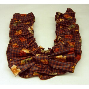 May include: A ruffled neck scarf featuring a dark brown and black gingham check pattern with an overlay of scattered autumn leaves in shades of orange, yellow, and green. The scarf is shaped into a U-form with a large, gathered bow at the center.