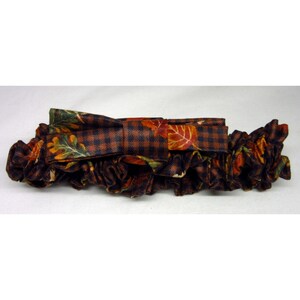 May include: A fabric hair scrunchie featuring a dark brown and black gingham check pattern overlaid with a colorful autumn leaf print. The accessory includes a central bow detail made from the same patterned fabric, creating a gathered, ruffled look.