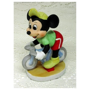 Puede incluir: Una figura de cer&aacute;mica de Mickey Mouse montando en bicicleta azul claro. Mickey lleva una camisa verde, pantalones cortos rojos, zapatos y gorra amarillos y guantes blancos. La figura est&aacute; sobre una base beige con un acento rojo.