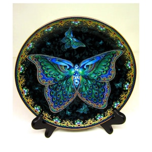 May include: Decorative plate featuring a large butterfly design with blue and green wings, set against a dark background. The plate has a gold rim with floral accents and is displayed on a black stand. The butterfly design is detailed and vibrant.