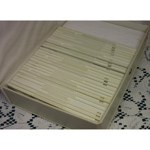 May include: An open, off-white storage case filled with numerous, thin, cream-colored plastic dividers. The dividers are arranged neatly inside the case, which has a light-colored interior and a simple, rectangular design. A white, padded material is visible at the top.