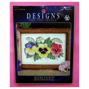 May include: A cross-stitch kit featuring a framed floral design of pansies in yellow, purple, and red. The kit is labeled "DESIGNS FOR THE NEEDLE" and "HOMETOWN". The package dimensions are 5" x 7" (12.7 x 17.8 cm).