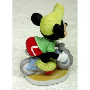 Puede incluir: Figura de cer&aacute;mica de Mickey Mouse en bicicleta. Mickey viste camisa verde, pantal&oacute;n corto rojo y gorra amarilla. La bicicleta es azul claro con ruedas grises. La figura est&aacute; sobre una base blanca con borde beige.